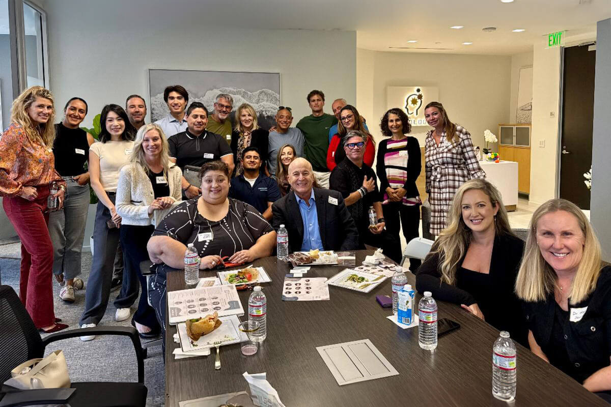 A large group of smiling people gathered around a conference table, enjoying food and conversation in a bright, modern office space. The atmosphere feels collaborative and friendly.