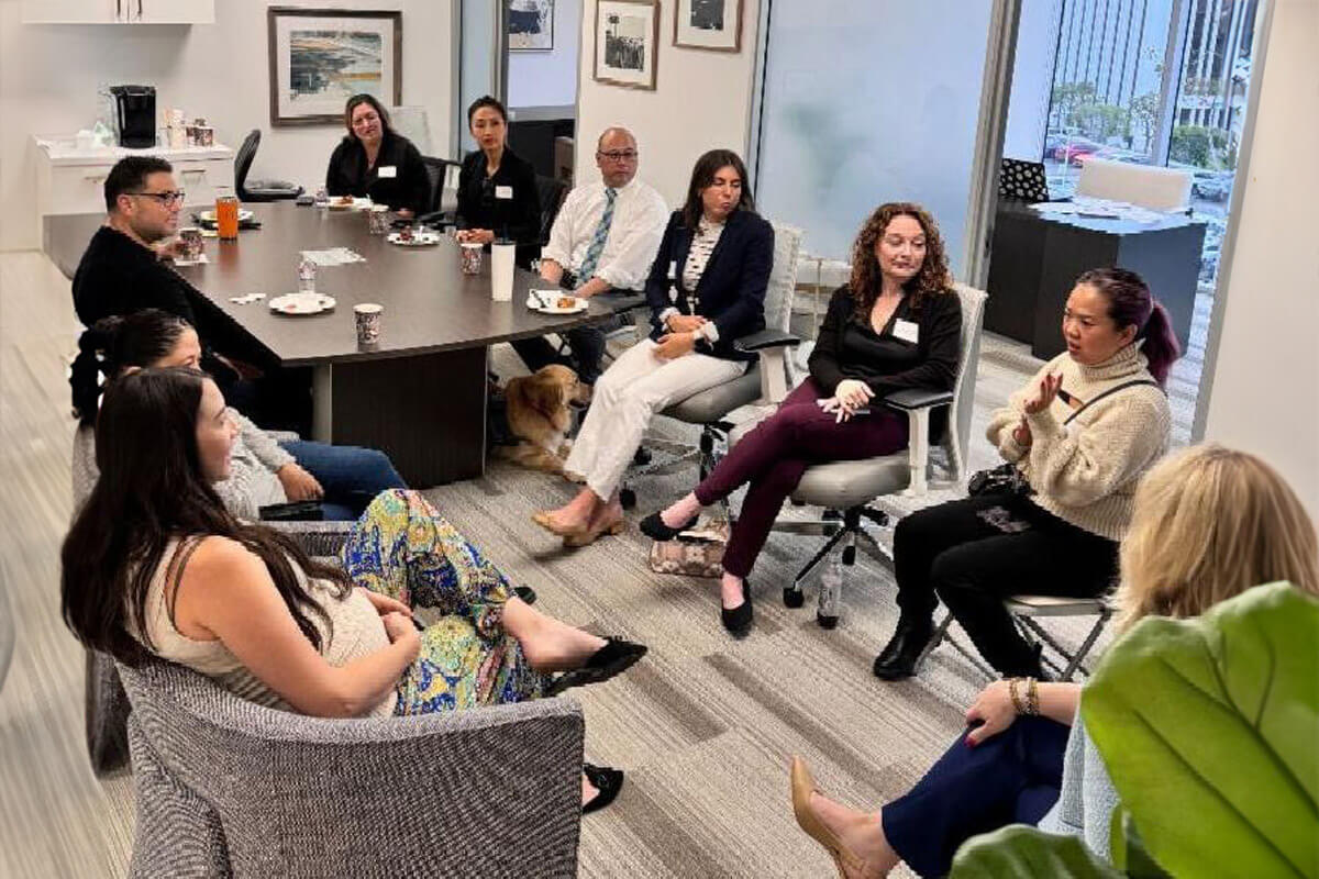 A small group meeting where participants sit around a conference table, attentively listening to a woman speaking. The mood is warm and inclusive, suggesting open dialogue and teamwork.