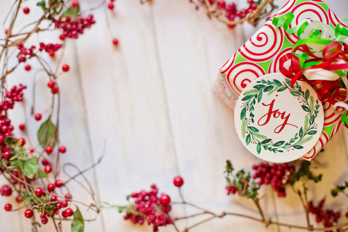 december_newsletter_2025_0 A festively wrapped holiday gift with a “Joy” tag sits surrounded by red berries on a light wooden surface.