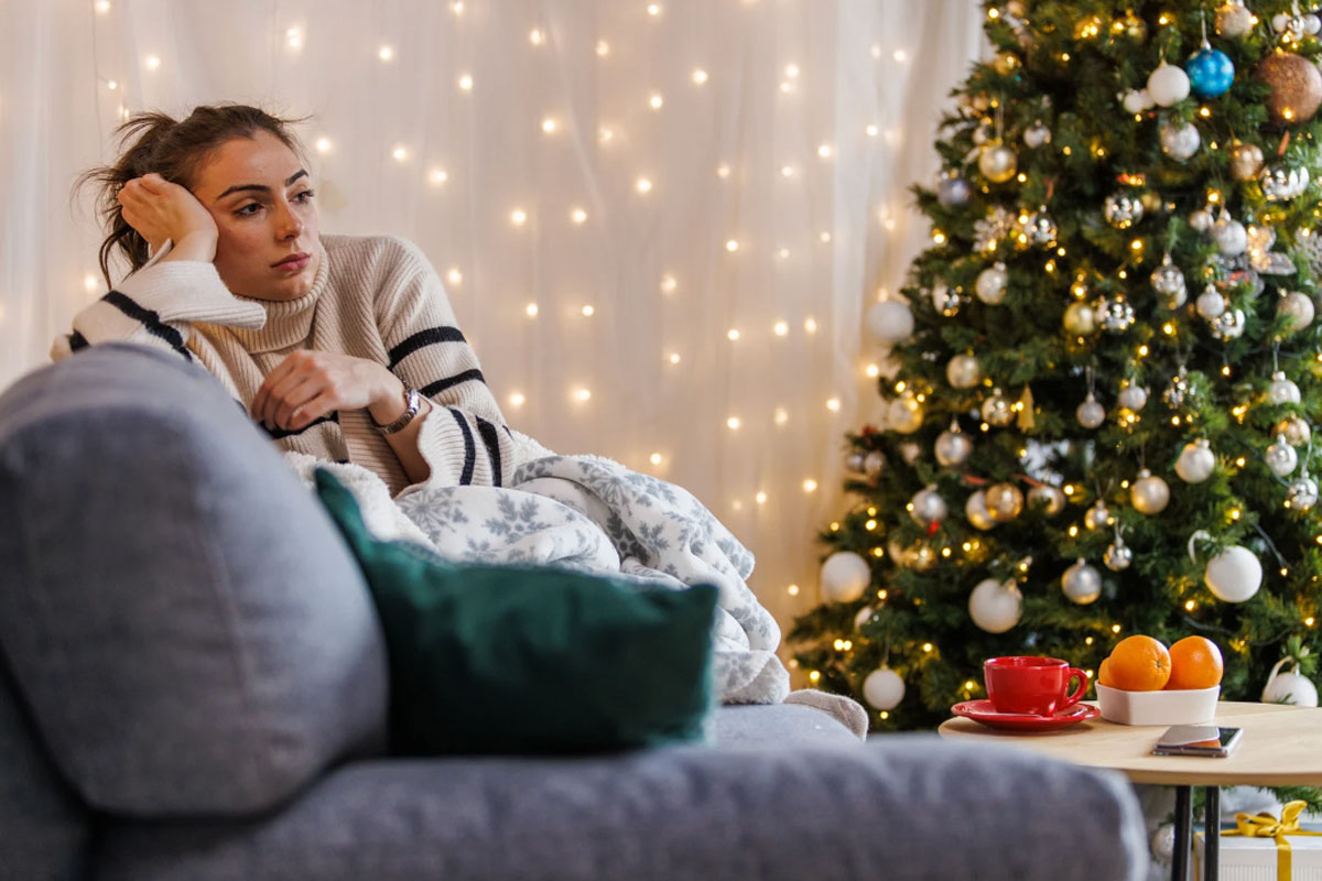 december_newsletter_2025_13 A woman sits on a couch looking sad or reflective beside a decorated Christmas tree and warm holiday lights.