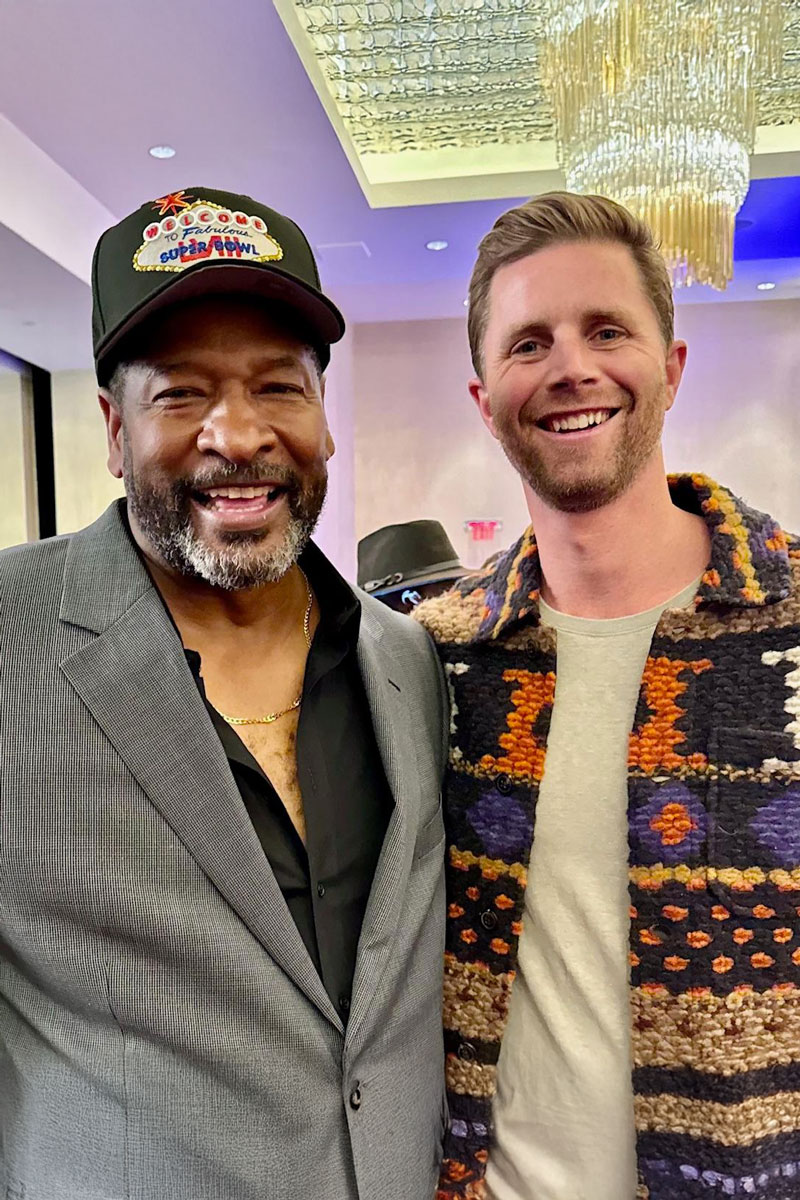 december_newsletter_2025_5 Two men smile for a photo at an indoor event, one wearing a Super Bowl–themed hat and the other a colorful patterned jacket.