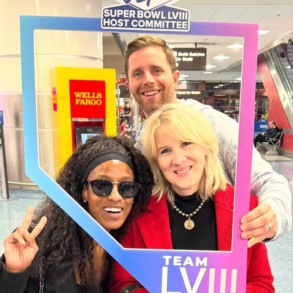 december_newsletter_2025_6 Three people pose inside an airport holding a Team LVIII Super Bowl host committee frame and smiling brightly.