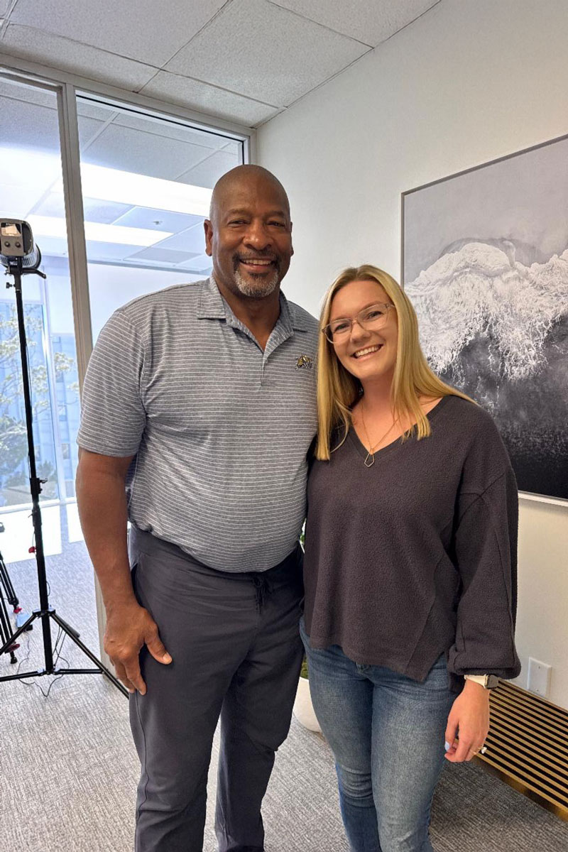 december_newsletter_2025_8 A man and a woman smile together in an office setting with professional lighting equipment behind them.