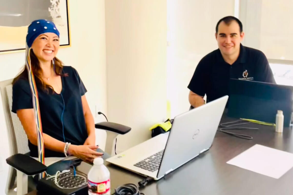 A woman wearing a brainwave cap sits in a clinical setting during a neurological assessment while a staff member monitors the session from a laptop.