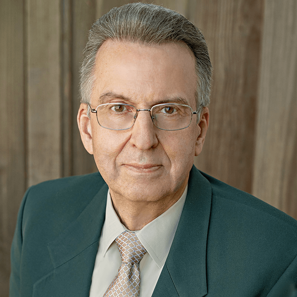 A professional headshot of an older man wearing glasses and a suit, posed against a neutral wood background.