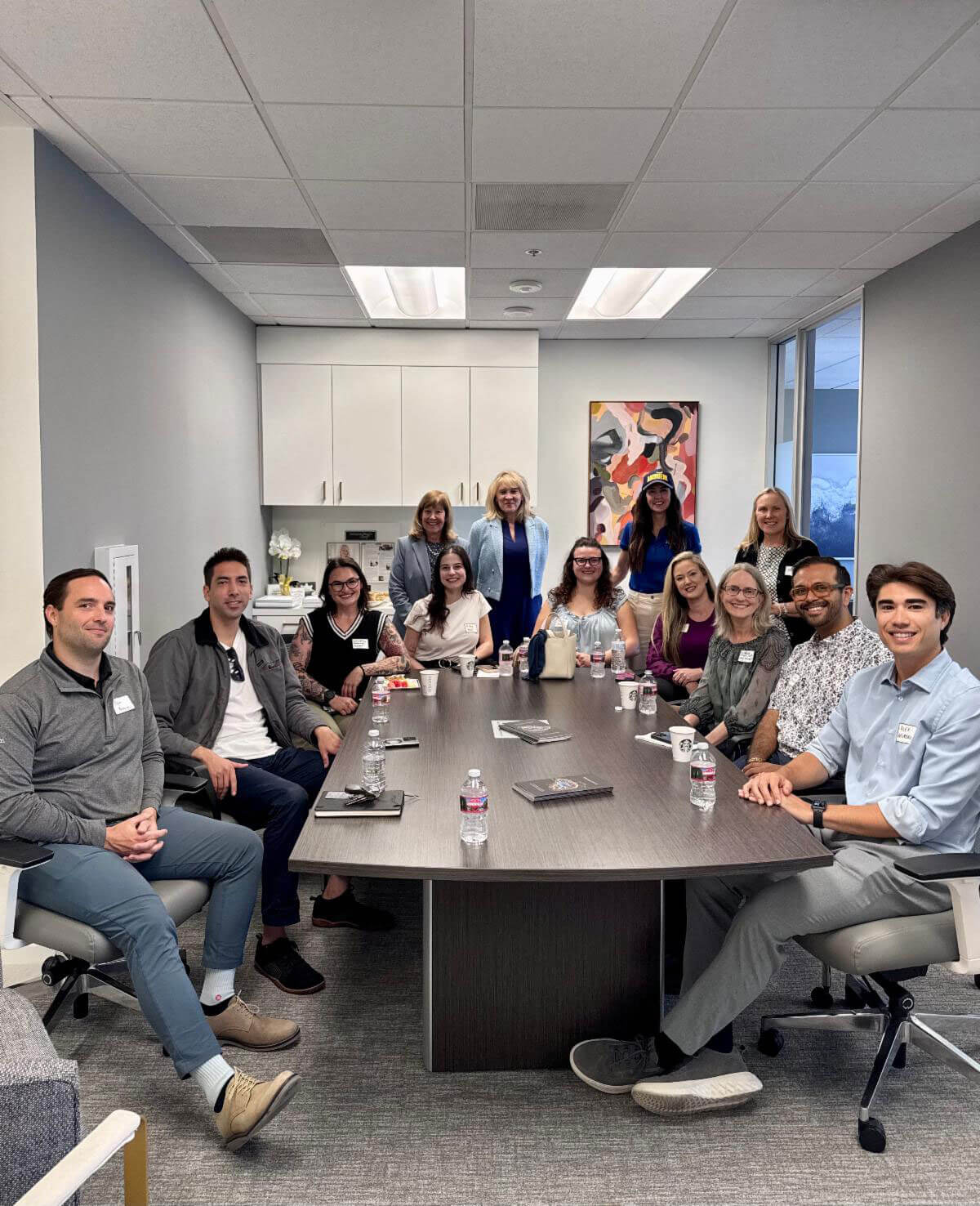april_newsletter_2026_4 A group of approximately fourteen smiling professionals wearing name tags gather around a large conference table with water bottles and notebooks in a bright modern office.