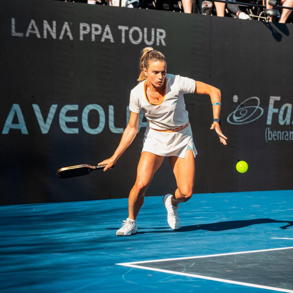 april_newsletter_2026_5 A female pickleball player in white athletic wear lunges forward to return a shot during a professional PPA Tour match on a blue outdoor court.