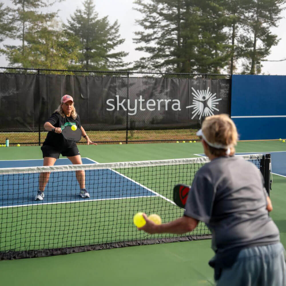 april_newsletter_2026_6 Two women practice pickleball across a net on an outdoor court with a Skyterra-branded fence banner and tall trees in the misty background.