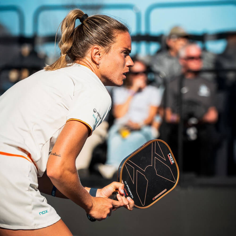 april_newsletter_2026_7 A focused female pickleball player in white and orange athletic wear holds a Nox paddle in a ready stance, photographed in close profile with a blurred spectator crowd behind her.
