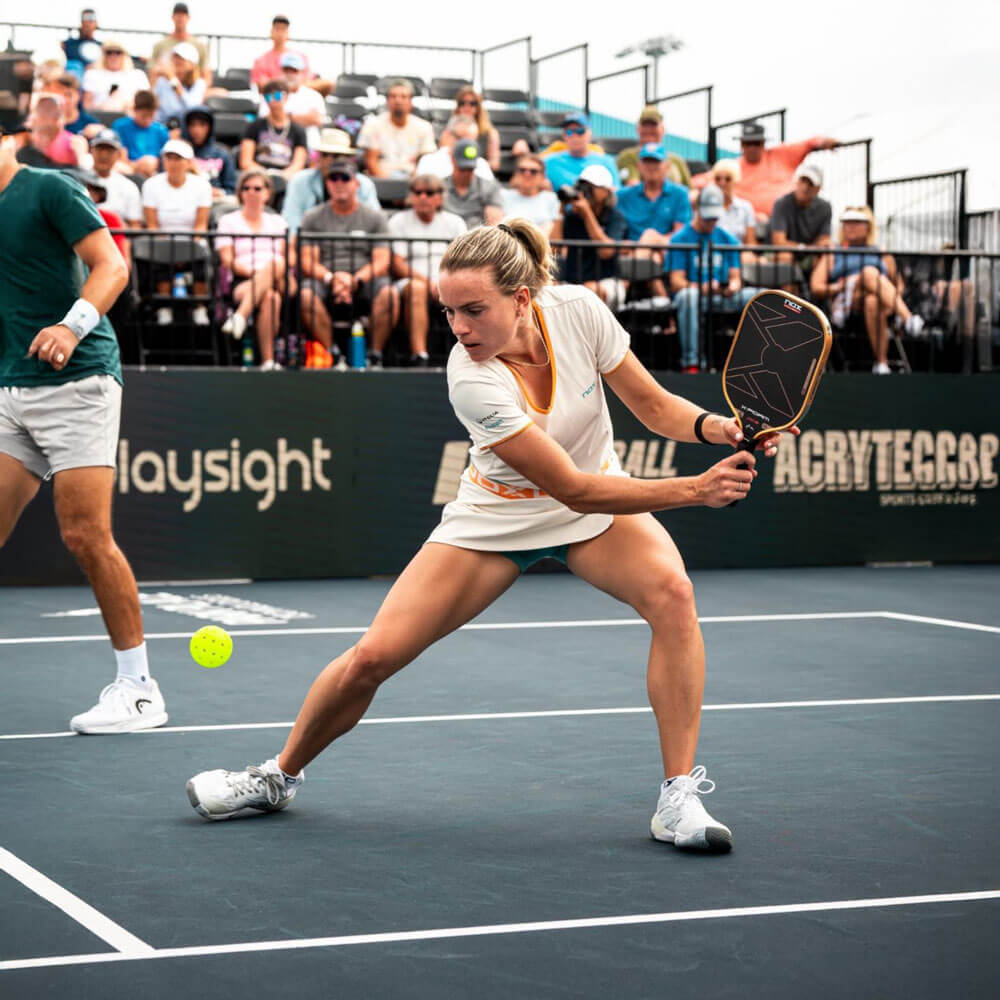 april_newsletter_2026_8 A female pickleball player in white stretches low to reach a ball during a professional doubles match on a dark court, with a large seated crowd visible in the stands.
