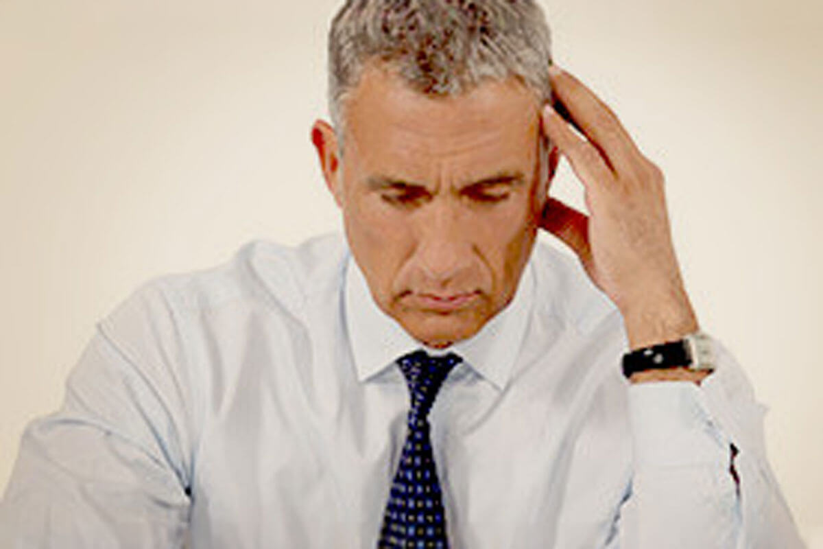 A middle-aged man in a shirt and tie looks down with a furrowed brow, pressing his hand to his temple as if experiencing stress or a headache.