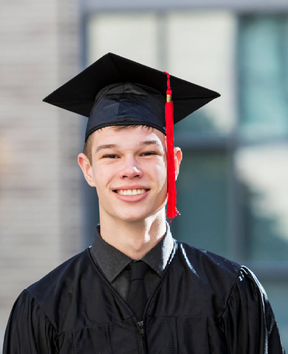 A smiling young man in a black graduation gown and cap with a red tassel poses outdoors in bright sunlight.