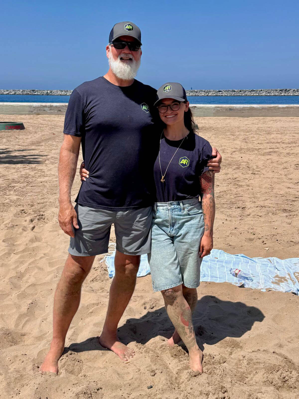 A man with a white beard and a smiling woman stand together on a sandy beach wearing matching dark t-shirts and gray baseball caps featuring an "AR" logo.