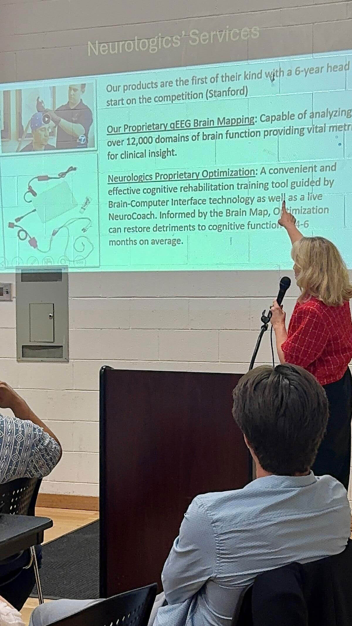 A woman holding a microphone points to a projected presentation slide detailing Neurologics' proprietary qEEG Brain Mapping and Optimization services while addressing an audience.