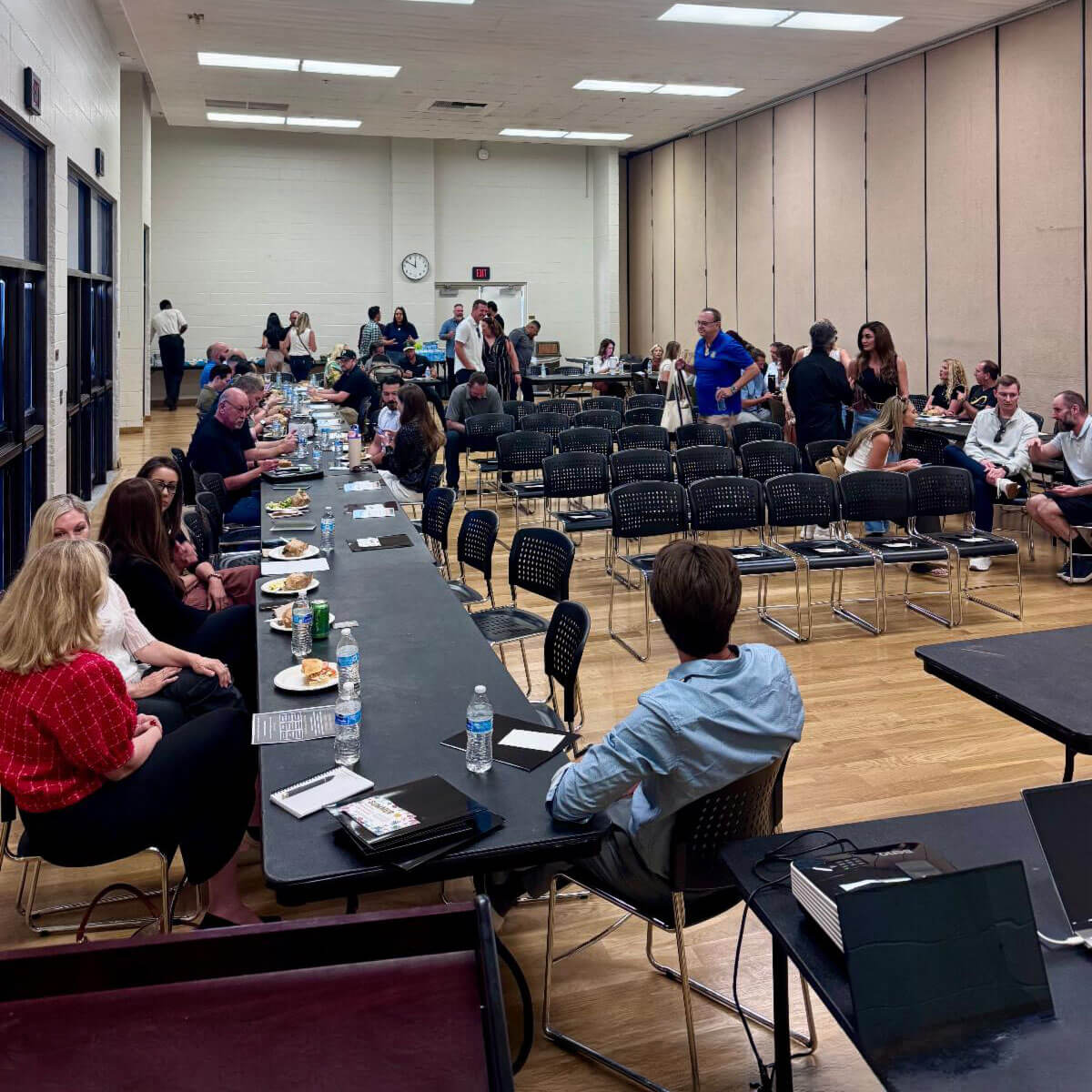 Dozens of attendees sit at long tables eating a catered meal and socializing in a spacious, brightly lit hall during a community event.