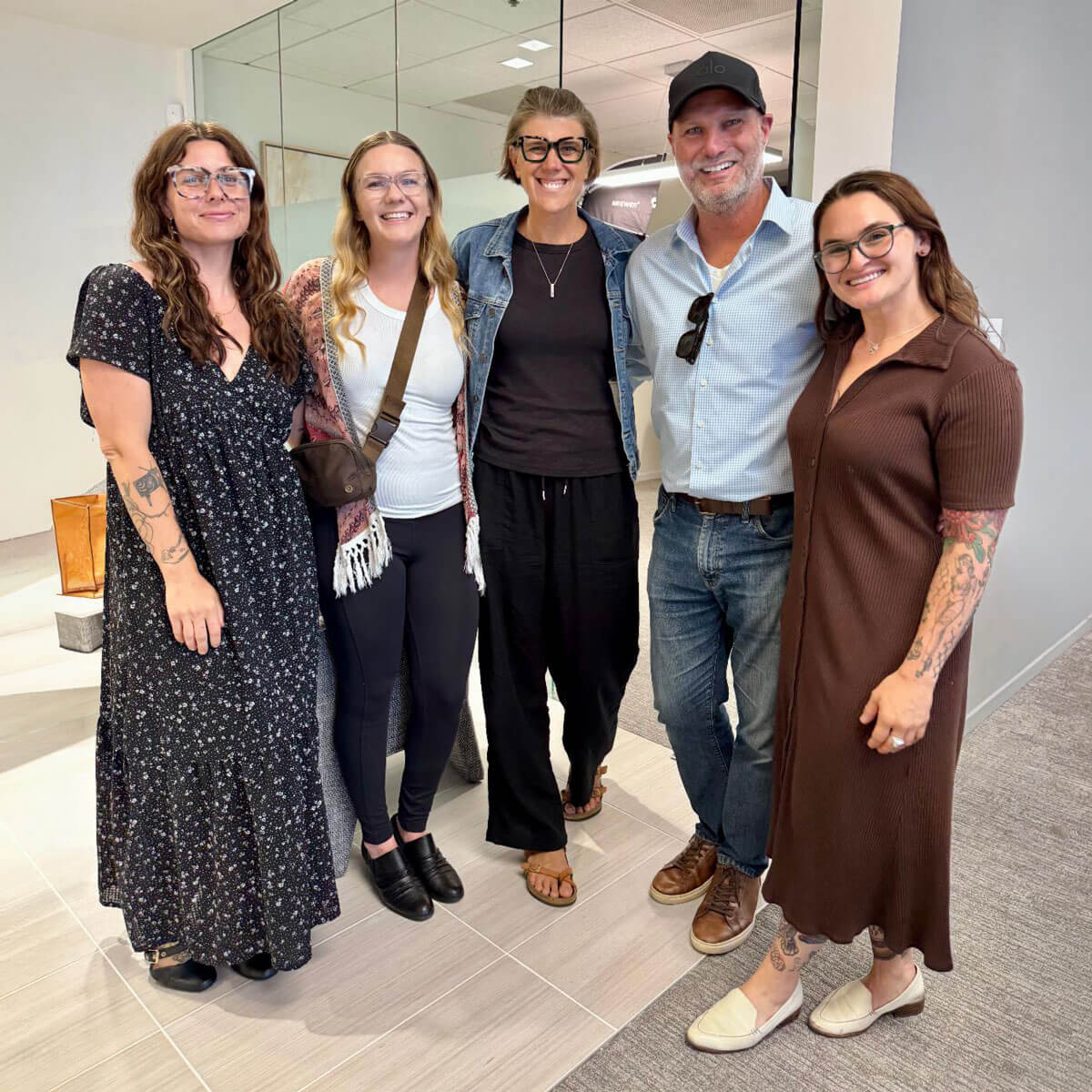 Five casually dressed professionals standing together and smiling warmly for a photo in a modern office hallway.