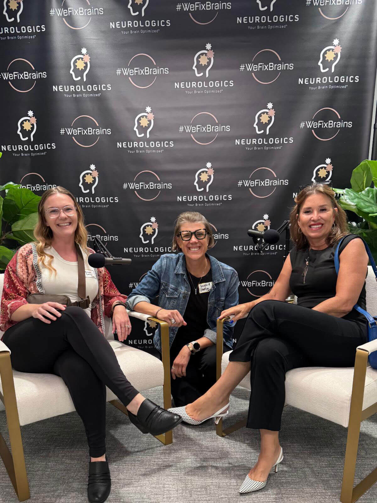 Three smiling women sit comfortably in armchairs around a microphone in front of a Neurologics step-and-repeat banner.