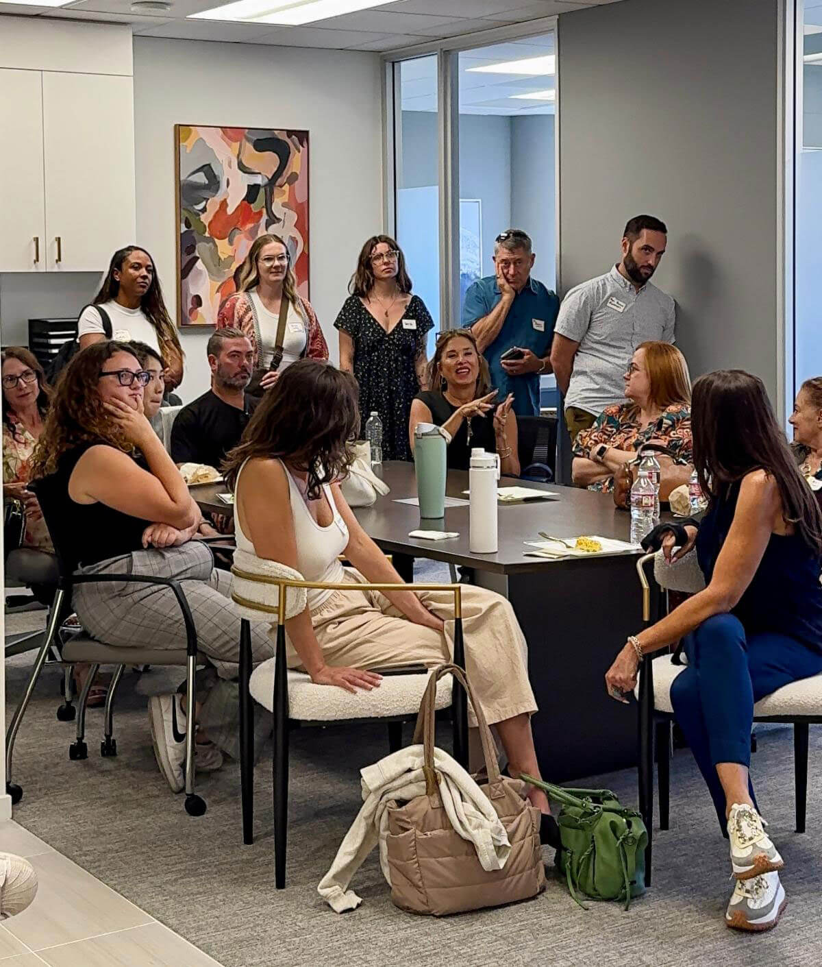 A diverse group of event attendees sit and stand around a conference room table, listening attentively to an off-camera speaker.