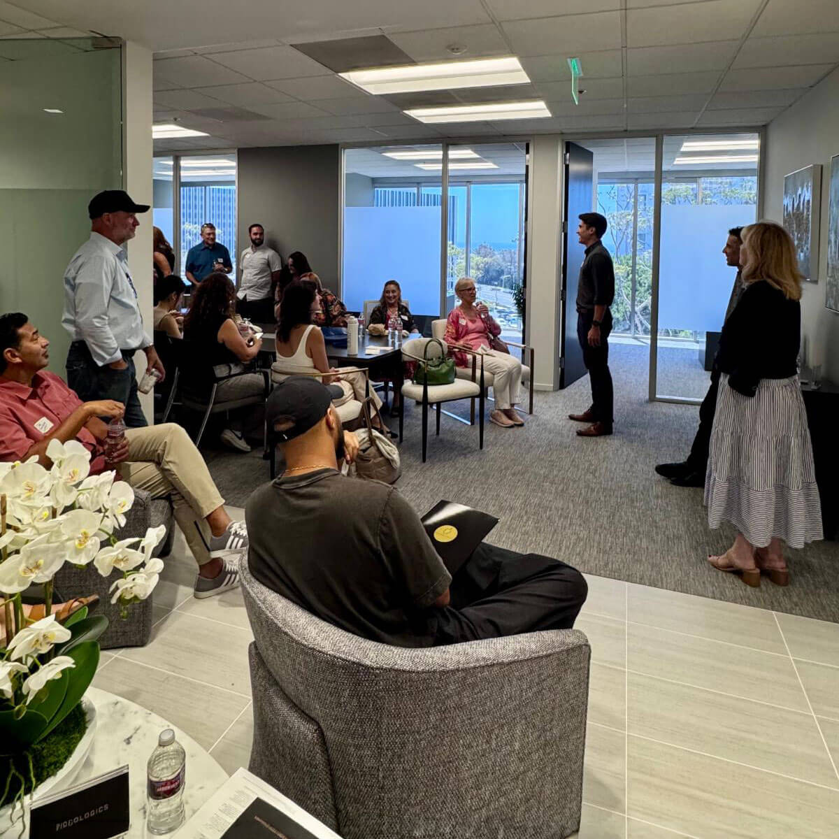 A male speaker stands and addresses an engaged group of attendees gathered in a bright, modern office lounge area.